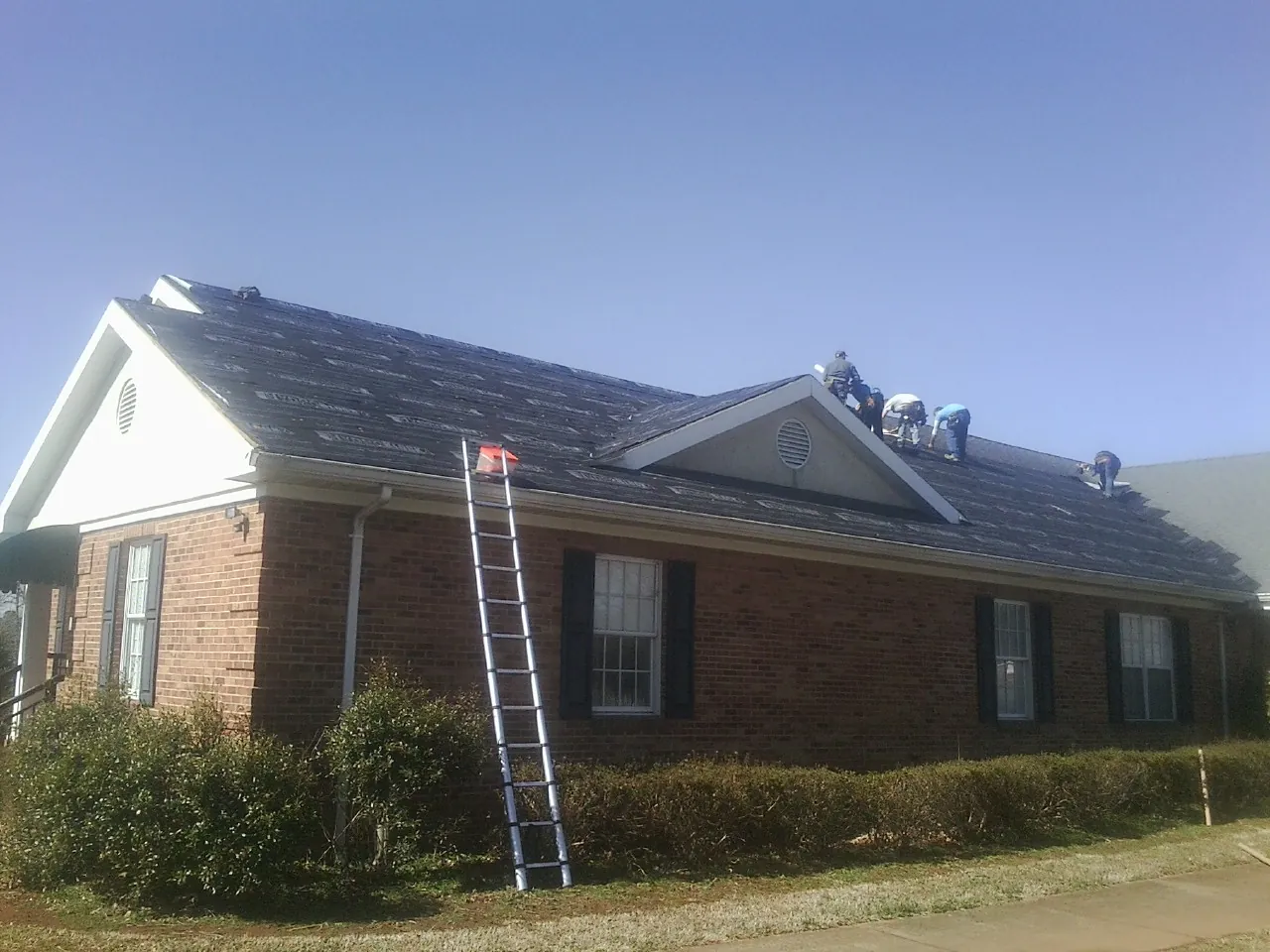 Crew tearing shingles off a brick ranch roof with a ladder set — Emergency Roof Repair work by our crew in Lynd
