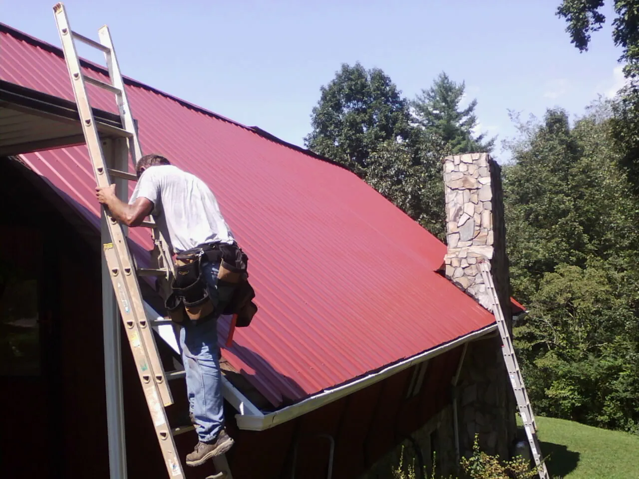Roofer on a ladder beside a red metal roof and stone chimney — Soffit & Fascia Repair by Roofing Brothers in Lynd